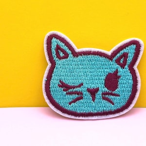 Könnte beinhalten: Gestickter Aufnäher mit einem türkisfarbenen Katzenkopf mit zwinkerndem Auge und weinroten Akzenten. Der Aufnäher hat einen weißen Rand und ist vor einem gelben Hintergrund platziert.