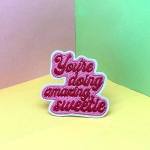 Könnte beinhalten: Gestickter Aufnäher mit den Worten "You're doing amazing sweetie" in rosa und roter Farbe. Der Aufnäher hat einen weißen Rand und ist vor einem bunten Hintergrund aus Rosa, Gelb und Grün platziert.