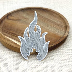 May include: A silver-grey embroidered patch in the shape of a stylised flame. The design features a dense, textured thread pattern with a clean white border outlining the jagged, upward-reaching curves of the fire motif, creating a distinct shape.