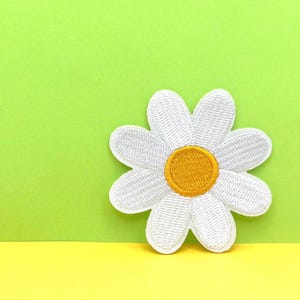 May include: A white daisy flower with a yellow center, embroidered on a green background.