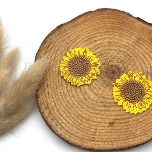 Puede incluir: Dos parches de girasol amarillos y marrones se muestran sobre una rodaja de madera redonda. Los girasoles tienen costuras detalladas. Hierba decorativa beige seca está a la izquierda. El fondo es blanco.
