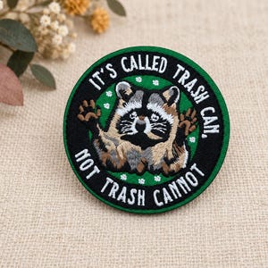 Könnte beinhalten: Ein runder gestickter Aufnäher mit einem braun-grauen Waschbären mit erhobenen Pfoten auf grünem Hintergrund. Der schwarze Rand enthält den weißen gestickten Text: IT'S CALLED TRASH CAN, NOT TRASH CANNOT, verziert mit kleinen weißen Blumen.