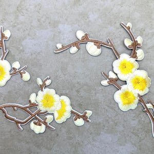 Puede incluir: Tres parches bordados que muestran ramas marrones con pétalos de flores blancos y centros de color amarillo brillante. Los diseños florales incluyen múltiples flores y pequeños capullos dispuestos en tallos curvos con un borde blanco.