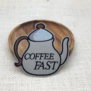 May include: An embroidered patch shaped like a teapot with a light gray body, a dark brown handle, and a light blue lid. The text COFFEE FAST is stitched in black capital letters across the center of the teapot body with a dark outline.