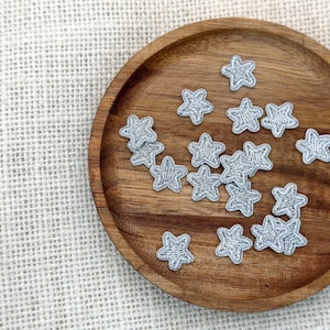 以下が含まれることがあります： 丸い木製のプレートに配置された、小さな銀色の星型の刺繍パッチのコレクション。パッチは詳細で質感のある外観で、プレートの表面に散らばっています。木製のプレートは豊かで暖かい色調です。