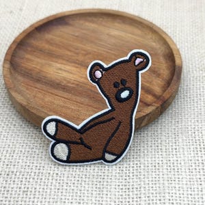 Puede incluir: Parche de oso de peluche marrón con detalles en negro y blanco. El oso está sentado con los brazos y las piernas extendidos. El parche está sobre una superficie beige claro con un plato de madera en el fondo.