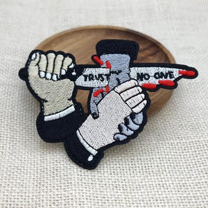 May include: An embroidered patch featuring two hands in black suit sleeves. One hand holds a white knife with red blood splatters and the text TRUST NO ONE, while the other hand grips the blade. The design uses black, white, grey, and red thread.