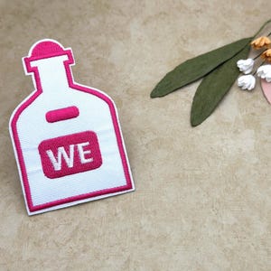 Puede incluir: Un parche blanco con forma de botella con contorno y detalles rosas. El parche tiene las letras "WE" en un rectángulo rosa. El fondo es de color beige neutro con hojas verdes y pequeñas flores blancas.