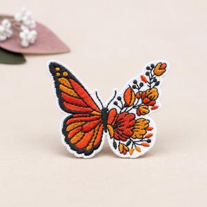 May include: An embroidered patch shaped like a butterfly with one wing featuring orange and black monarch patterns and the other wing composed of orange, yellow, and black floral and botanical elements with a white border outline.