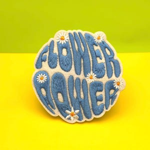 Pode incluir: Patch bordado com as palavras "FLOWER POWER" em escrita azul sobre um fundo branco. Pequenos detalhes de margaridas são costurados nas bordas. O patch está sobre um fundo amarelo e verde.