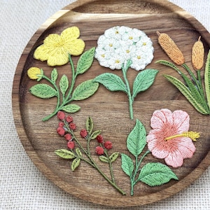 May include: A collection of five embroidered floral and botanical patches arranged on a round wooden tray. The set includes a yellow flower, a white hydrangea, two brown cattails, a sprig of red berries, and a pink hibiscus flower with green leaves.