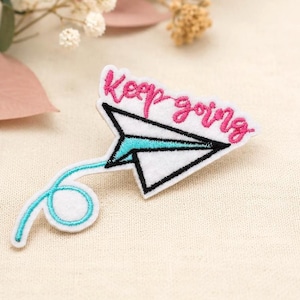 May include: An embroidered patch featuring a white paper airplane with a black outline and a bright turquoise trail. Above the airplane, the text "Keep going" is stitched in vibrant pink thread. The patch has a white felt base.