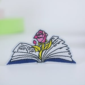 May include: An embroidered iron-on patch featuring a pink rose with a yellow stem and leaves emerging from an open book with blue pages.