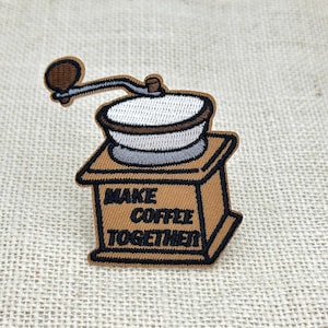 Könnte beinhalten: Ein gestickter Aufnäher, der eine braune Kaffeemühle mit weißem Oberteil und schwarzem Griff zeigt. Der Text "MAKE COFFEE TOGETHER" ist in schwarzen Buchstaben auf der Vorderseite des quadratischen Sockels der Kaffeemühle eingestickt.