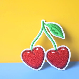 Puede incluir: Parche bordado con dos cerezas rojas en forma de corazón con contornos blancos, tallos verdes y una hoja. El parche está sobre un fondo amarillo y azul, creando un contraste vibrante.