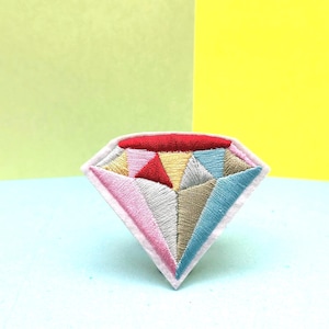 May include: A colorful embroidered diamond-shaped patch with red, yellow, blue, pink, and white thread. The patch is on a light blue surface.