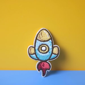 May include: A blue and yellow embroidered rocket patch with a red flame. The rocket has a white window and is surrounded by a white outline.
