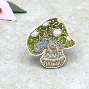May include: Embroidered mushroom patch with a green and white cap, gray stem, and gold accents. The patch features a crescent moon and star design. The patch is on a white background.