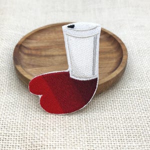 May include: An embroidered patch shaped like a glass of red wine. The design features a white cylindrical glass with a dark red liquid base, rendered in detailed thread work with a visible texture and a distinct colour gradient from bright to deep red.