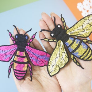 May include: Two embroidered bee patches, one with yellow and blue wings and the other with pink and purple wings. Both bees have black bodies with yellow and gold stripes.