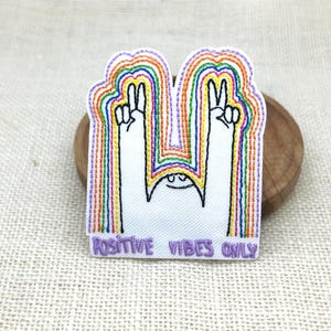Puede incluir: Un parche bordado que muestra una figura blanca con dos brazos levantados haciendo signos de paz. La figura está rodeada por arcos concéntricos de colores del arcoíris en rojo, naranja, amarillo, verde, azul y morado. El texto POSITIVE VIBES ONLY está bordado en morado en la parte inferior.