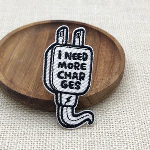 Könnte beinhalten: Ein schwarz-weiß gestickter Aufnäher in Form eines Steckers. Der Aufnäher trägt den Text "I NEED MORE CHARGES". Der Aufnäher liegt auf einem Holztablett.