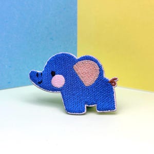 Op de afbeelding: Geborduurde blauwe olifantenpatch met roze oor- en wangdetails. De patch heeft een lachend gezicht en een kleine staart. De patch is waarschijnlijk voor kleding of accessoires.