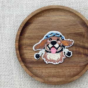 May include: Embroidered patch featuring a smiling dog wearing a blue aviator cap. The dog has brown fur, floppy ears, and a pink tongue. The patch is on a round, wooden tray with a light-colored fabric background.