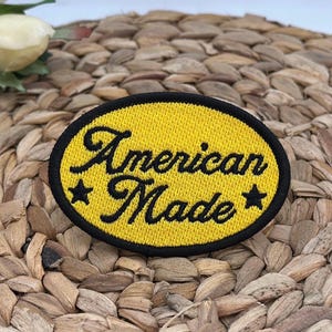 K&ouml;nnte beinhalten: Ein ovaler, bestickter Aufn&auml;her mit leuchtend gelbem Hintergrund und einem dicken schwarzen Rand. Der Schriftzug American Made ist in schwarzer Schreibschrift in der Mitte eingestickt und wird von einem kleinen schwarzen Stern flankiert.
