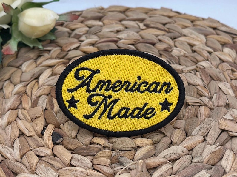 K&ouml;nnte beinhalten: Ein ovaler, bestickter Aufn&auml;her mit leuchtend gelbem Hintergrund und einem dicken schwarzen Rand. Der Schriftzug American Made ist in schwarzer Schreibschrift in der Mitte eingestickt und wird von einem kleinen schwarzen Stern flankiert.