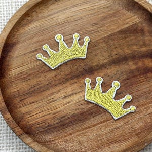 Puede incluir: Dos pequeños parches bordados en forma de corona amarilla con bordes blancos. Cada corona presenta cinco puntas rematadas con pequeños círculos, realizados con un patrón de hilo denso y texturizado que crea un aspecto metálico vibrante.