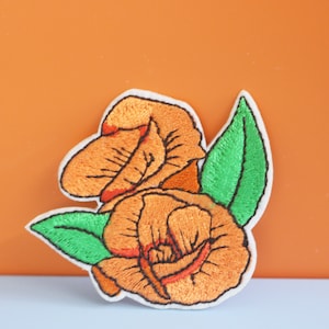 May include: Orange embroidered flower patch with green leaves. The patch is iron-on and can be used to decorate clothing, bags, or other items.