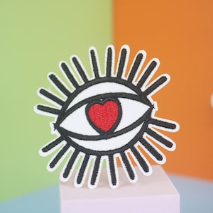 May include: A white embroidered patch with a black eye and a red heart in the center. The eye has black eyelashes and is surrounded by black rays.