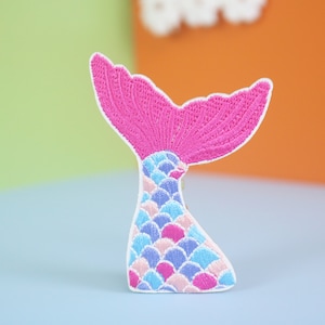 以下が含まれることがあります： 白い輪郭のあるピンクとブルーの刺繍された人魚の尾のパッチ。尾は鱗でできており、上にヒレがあります。