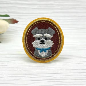 May include: Embroidered patch featuring a Schnauzer dog. The patch has a round shape with a yellow border and a brown background. The dog is gray and white with a blue bow tie.