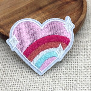 May include: Embroidered patch in the shape of a pink heart with a rainbow design and white stars. The patch has a light blue border and is likely for clothing or accessories.