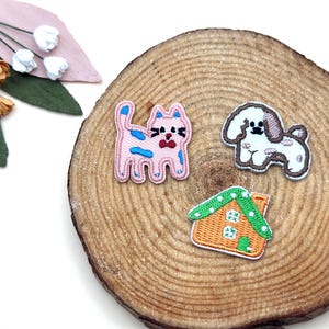 Può includere: Toppe ricamate raffiguranti un gatto rosa con dettagli blu, un cane bianco e marrone e una casa arancione con tetto verde. Le toppe sono su una fetta di legno con sfondo bianco.