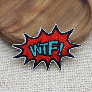 Könnte beinhalten: Ein bestickter Aufnäher im Comic-Stil mit einer gezackten, roten Sprechblasenform und einem dicken schwarzen Rand. Der Text WTF! ist in leuchtend cyanblauen Buchstaben in der Mitte des roten Hintergrunds gestickt, was ein kühnes Design ergibt.