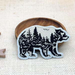 May include: An embroidered patch in the shape of a bear with a light cream background and black thread detailing. The bear's body features a detailed forest landscape with evergreen trees and small birds, finished with a light blue border edge.