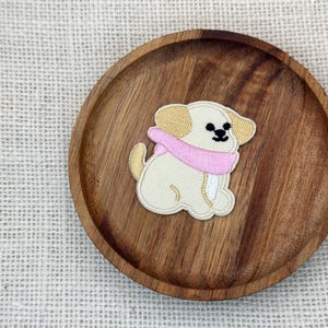 May include: An embroidered patch featuring a cream-colored puppy with tan ears and a pink scarf. The patch is sitting on a round, wooden plate with a natural wood grain pattern. The background is a light-colored woven fabric.
