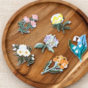 May include: Six embroidered flower patches featuring various botanical designs in shades of pink, yellow, white, purple, blue, and orange with green stems and leaves. The patches are arranged on a round wooden tray with a visible wood grain texture.