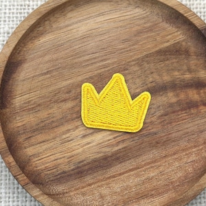 Puede incluir: Un parche de corona bordada amarilla. El parche tiene la forma de una corona clásica con cinco puntas. El parche está sobre un posavasos de madera con vetas visibles.