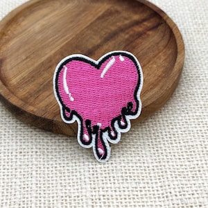 Puede incluir: Un parche bordado con forma de corazón con efecto de goteo. El corazón es de color rosa brillante con reflejos blancos y un contorno negro grueso. El bordado presenta puntadas densas en todo el diseño, creando una superficie texturizada.