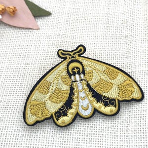 May include: Embroidered moth patch with gold and black detailing. The moth features intricate wing patterns and a black border. The patch is set against a textured, off-white backdrop. Decorative floral elements are visible in the upper left corner.