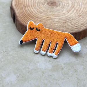 May include: Embroidered orange fox patch with white and black accents. The fox has a white-tipped tail, white paws, and closed eyes. This patch is suitable for clothing, bags, or other fabric items. The patch is approximately 3 inches long.