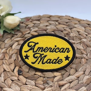 K&ouml;nnte beinhalten: Ein ovaler, bestickter Aufn&auml;her mit leuchtend gelbem Hintergrund und einem dicken schwarzen Rand. Der Text "American Made" ist in schwarzer Schreibschrift in der Mitte eingestickt, flankiert von einem kleinen schwarzen Stern auf beiden Seiten.