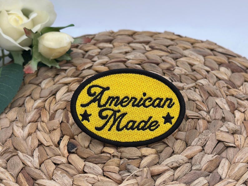 K&ouml;nnte beinhalten: Ein ovaler, bestickter Aufn&auml;her mit leuchtend gelbem Hintergrund und einem dicken schwarzen Rand. Der Text "American Made" ist in schwarzer Schreibschrift in der Mitte eingestickt, flankiert von einem kleinen schwarzen Stern auf beiden Seiten.
