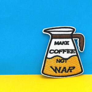 Puede incluir: Un parche bordado para planchar que representa una cafetera con el texto "Make Coffee Not War" en letras blancas y negras. La cafetera está llena de café amarillo y tiene un asa marrón.