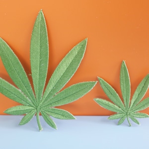 Pode incluir: Dois patches de folhas de feltro verde em forma de folhas de marijuana. A folha maior está à esquerda e a menor à direita. As folhas estão sobre uma superfície azul claro.
