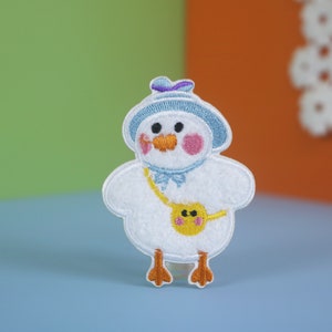 May include: A white embroidered patch of a cartoon chick wearing a blue hat and carrying a yellow bag. The chick has pink cheeks and a small orange beak.